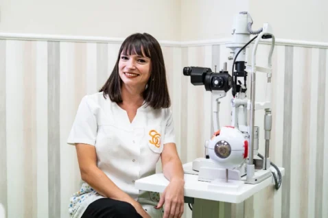Priscilla Viotto sonriendo en consulta junto a un modelo anatómico del ojo humano y equipos de diagnóstico oftalmológico.