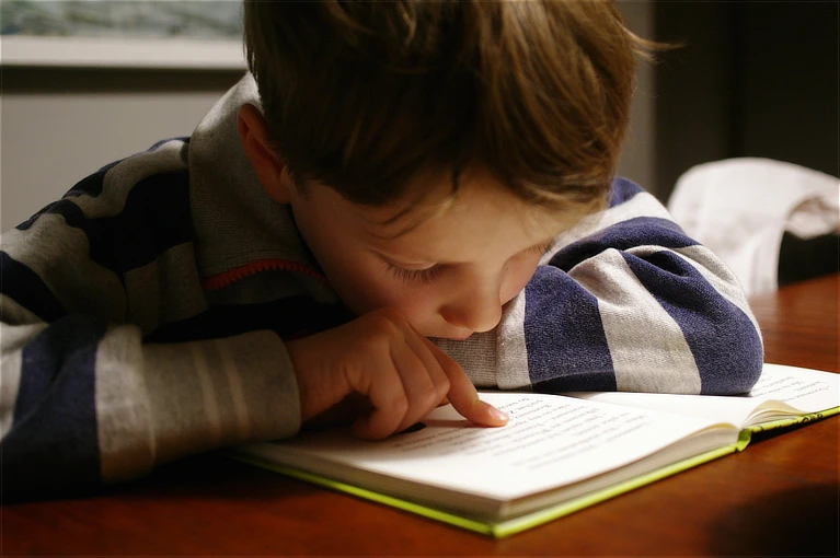 Niño inclinado muy cerca de un libro abierto, siguiendo la lectura con el dedo y mostrando una expresión de intenso esfuerzo y concentración bajo una luz cálida y tenue.